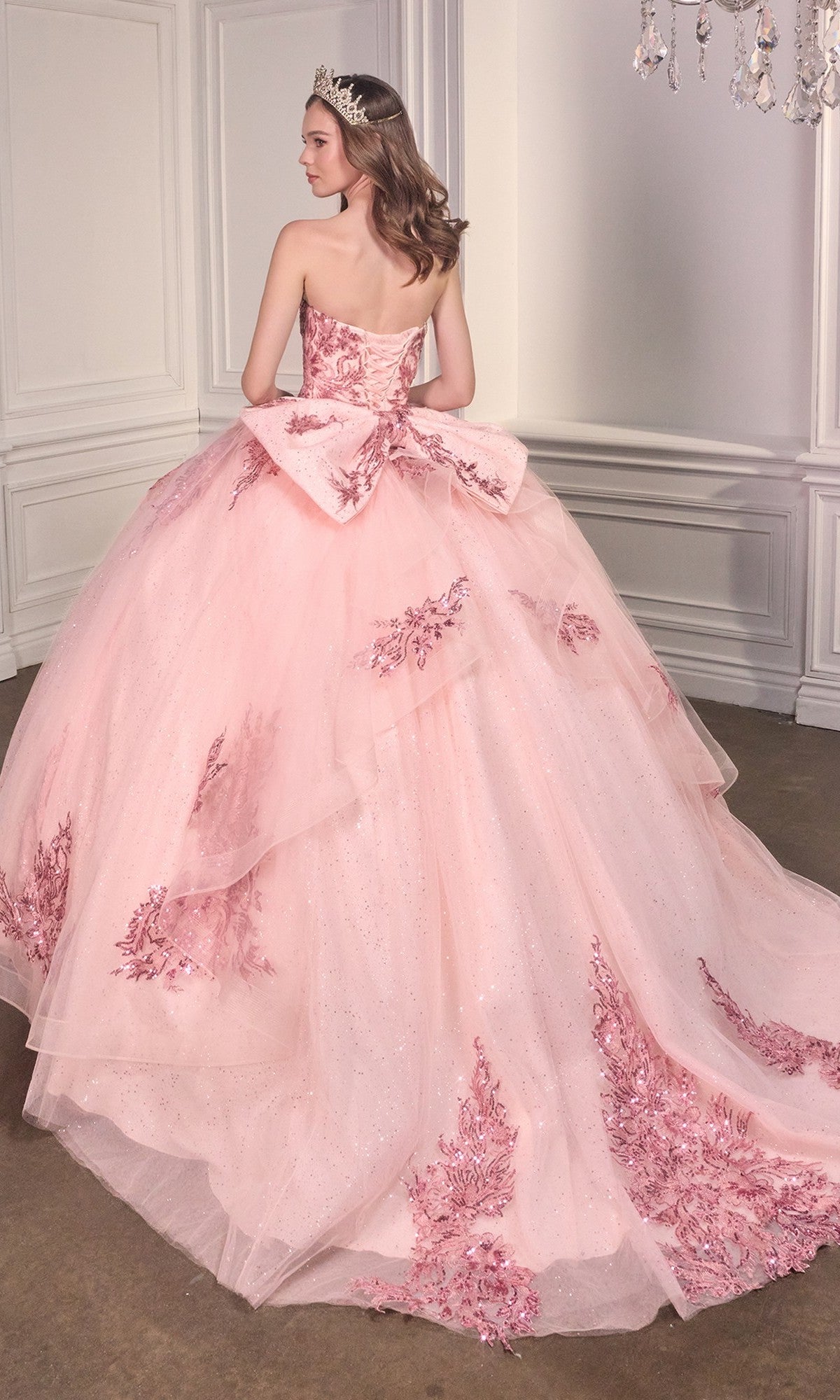 A woman wearing the Ladivine Strapless Quinceanera Dress 15715, adorned with glitter and floral appliqués, stands in an elegant room, facing away from the camera.